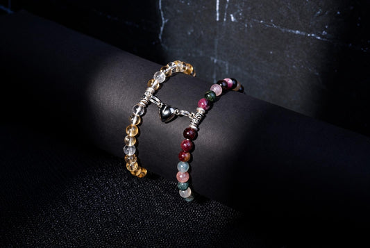 Two beaded bracelets displayed on a black cylindrical stand. One bracelet features translucent yellow and clear beads with a silver heart charm. The other has multicolored round beads in shades of red, pink, green, and blue.