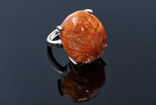 A silver-tone ring holds a polished oval stone filled with fine orange strands on a dark reflective surface.