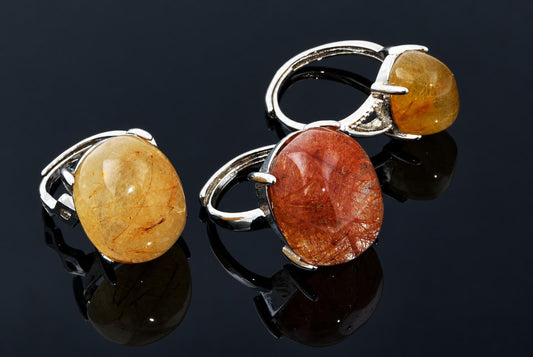 Silver-tone rings hold polished oval yellow and orange stones with fine internal strands on a dark reflective surface.