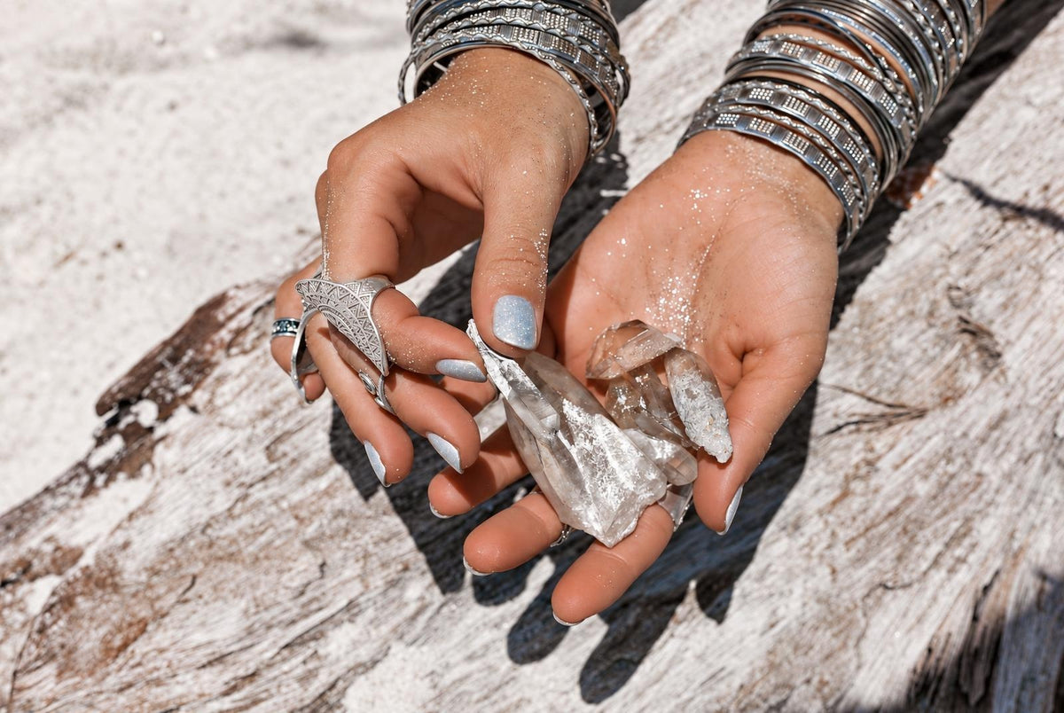 Hands wearing metallic rings and bangles hold clear rough crystals above a pale wooden surface.
