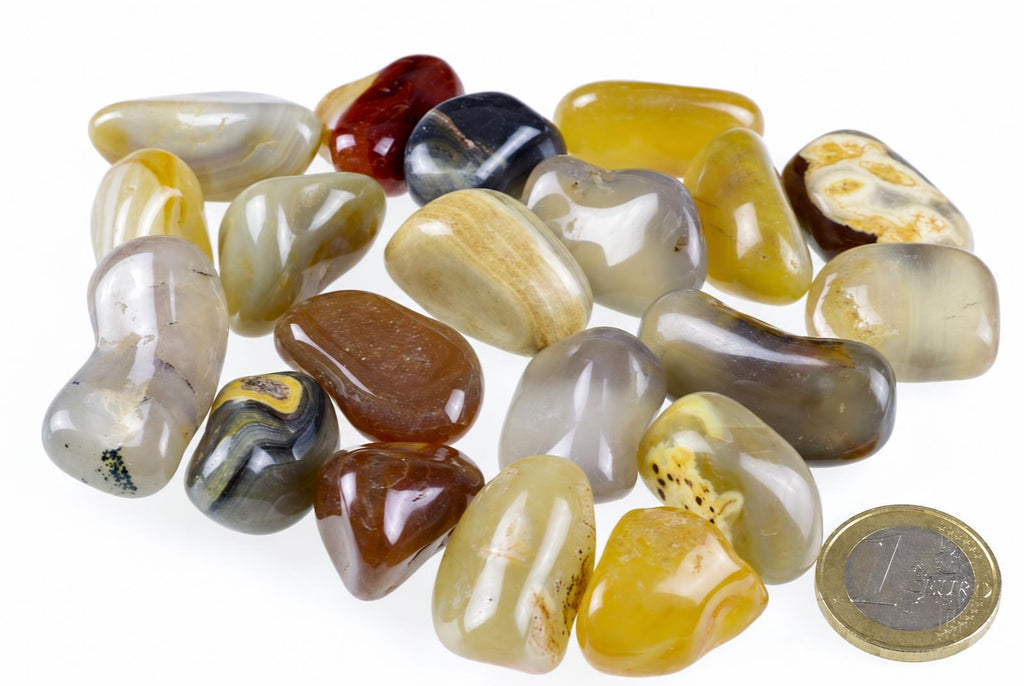 A collection of polished agate stones in various colours including yellow, brown, grey and black on a white surface with a one euro coin for scale.