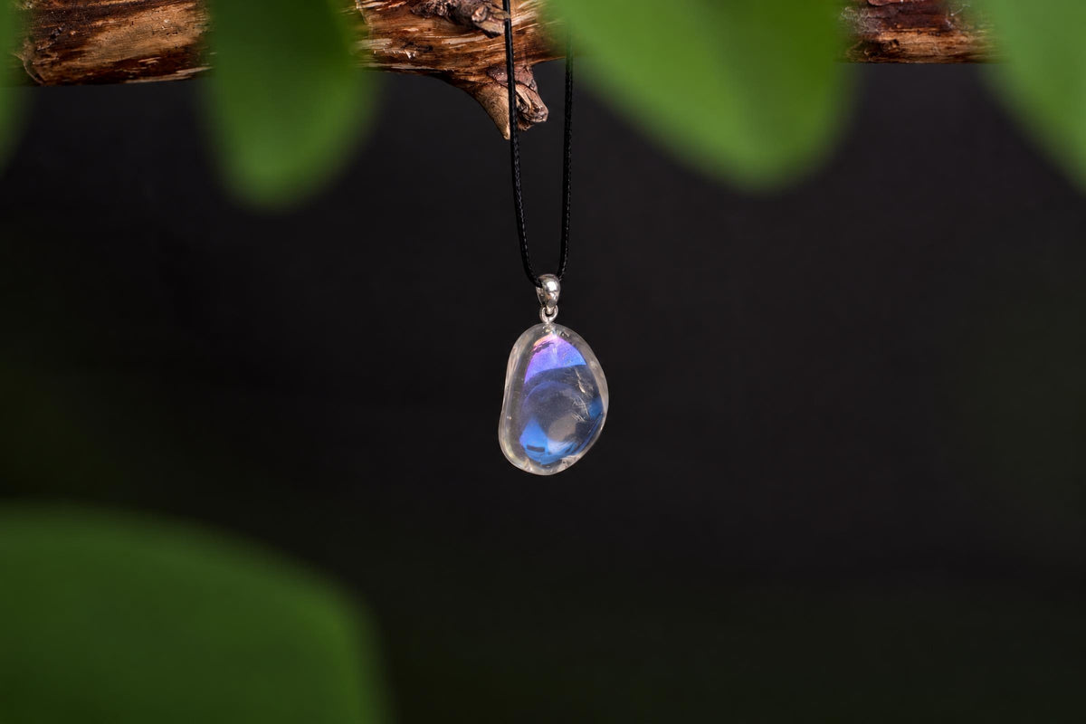 A teardrop-shaped pendant with a clear, iridescent stone showing blue and purple hues hangs from a black cord on a dark surface.