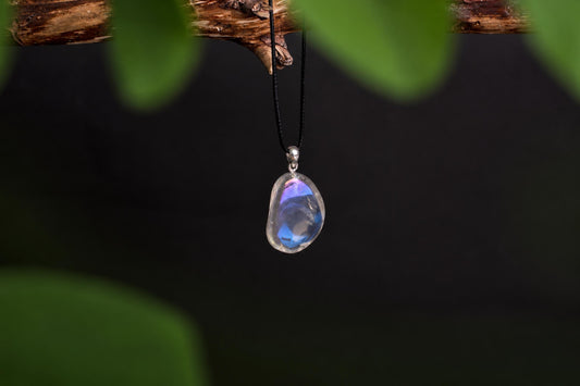 A teardrop-shaped pendant with a clear, iridescent stone showing blue and purple hues hangs from a black cord on a dark surface.