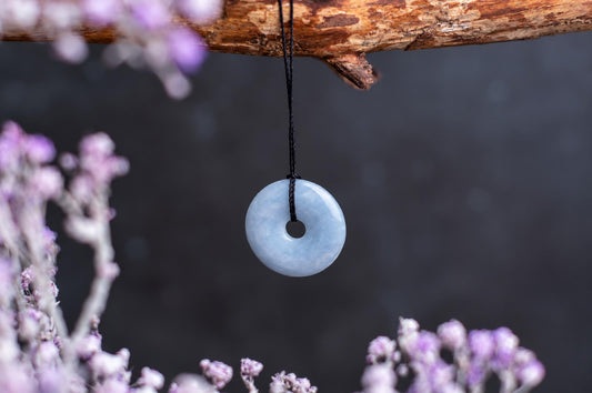 A light blue, donut-shaped pendant hangs from a black cord against a dark background with blurred purple flowers.