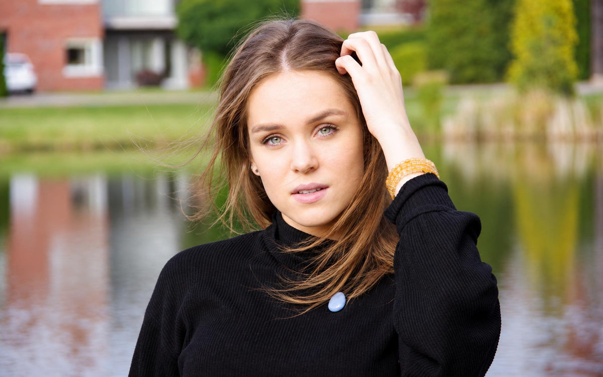 A woman wears a black turtleneck with a light blue oval pendant and yellow beaded bracelet, standing by water.