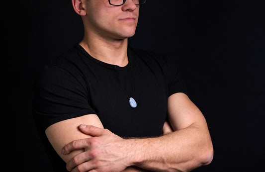 A man in a black t-shirt wears a light blue, smooth, oval pendant on a thin cord against a dark background.