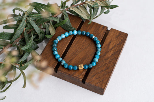 A blue beaded bracelet with a gold-toned square charm rests on a dark wooden surface beside green leaves.