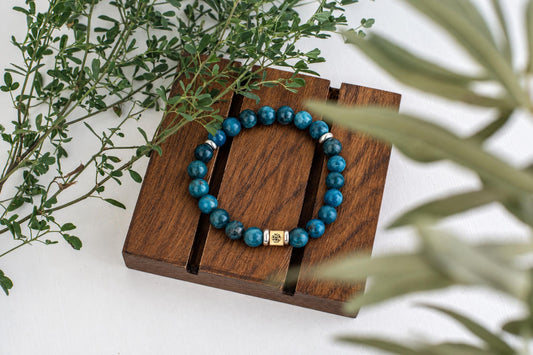 A blue beaded bracelet with silver-toned accents rests on a dark wooden surface, surrounded by green foliage.