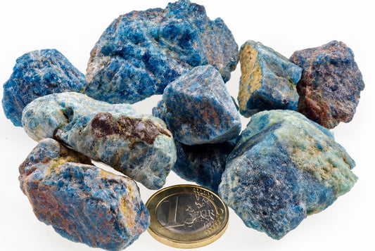 A pile of raw apatite crystals in blue, green and brown tones on a white surface with a one euro coin for scale.