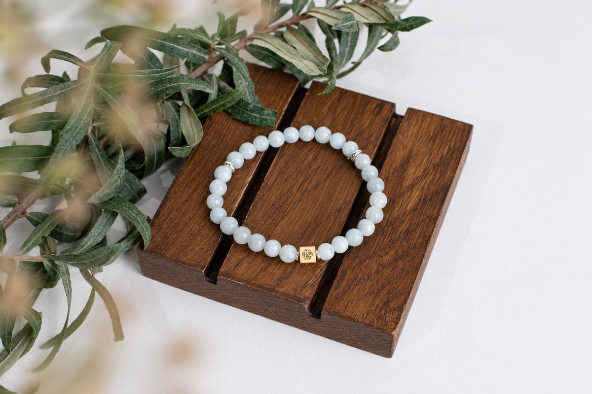 A bracelet of round pale blue beads with a gold-coloured accent bead rests on a wooden block beside leaves.