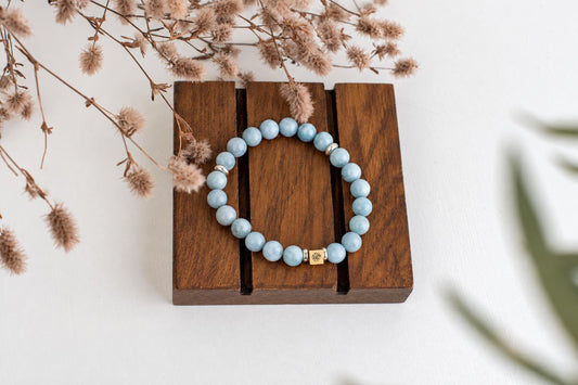 A bracelet with light blue beads and a gold-toned square charm rests on a dark wooden surface.