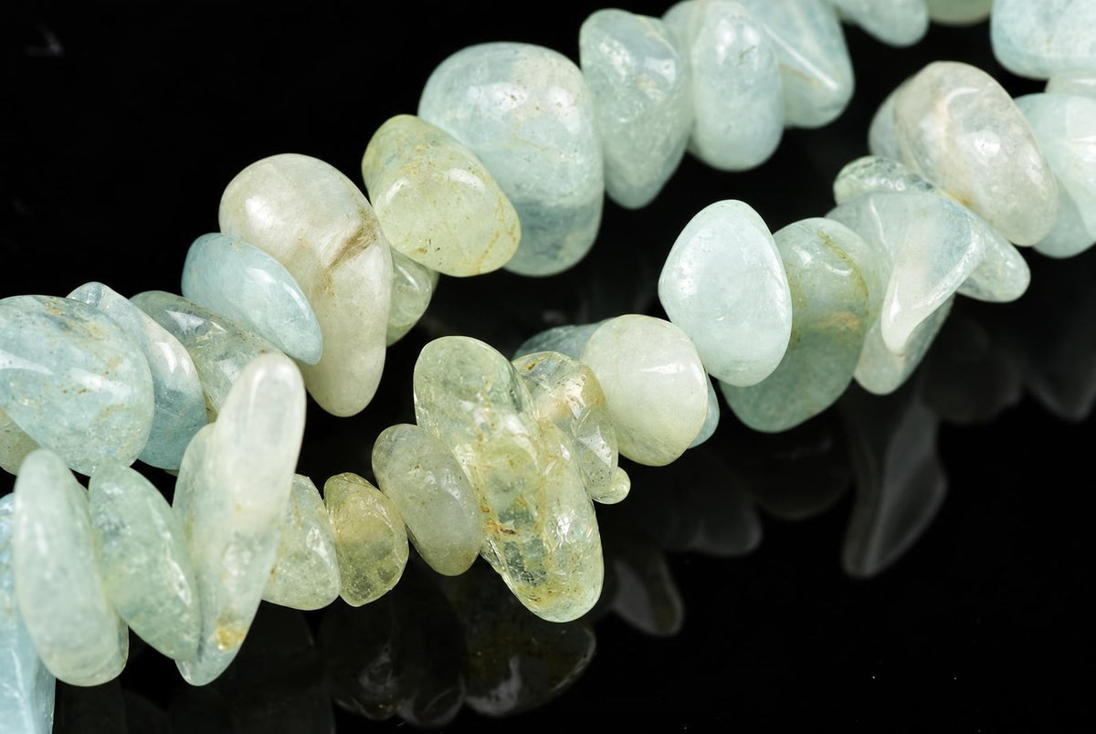 A strand of polished aquamarine chips in pale blue and green tones, arranged on a dark reflective surface.