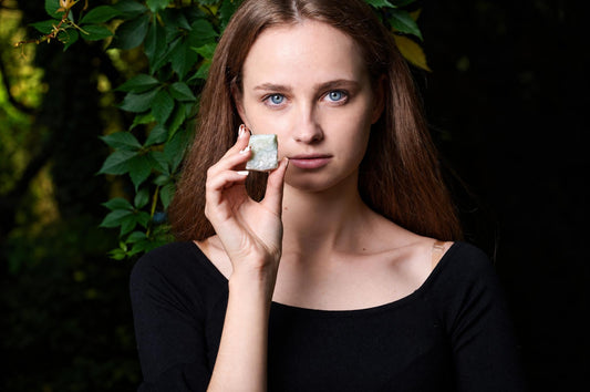 A woman holds a pale green, rough-textured stone near her face against a dark background with green leaves.