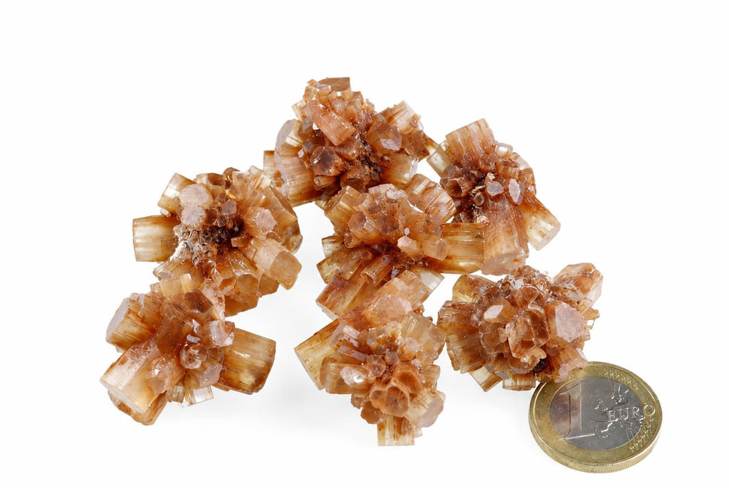 A cluster of brownish, translucent aragonite crystals with a hexagonal shape sits on a white surface next to a one-euro coin.