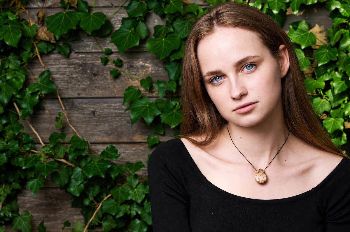 A woman wears a pendant with an oval, textured brown drop on a dark cord against a wooden wall with green ivy.