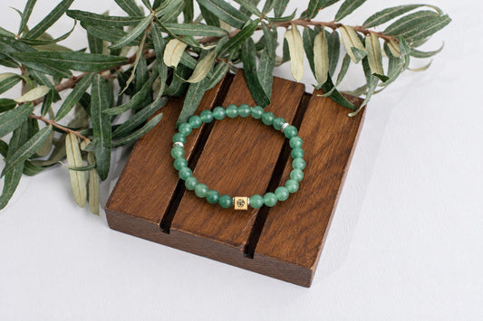 A bracelet with green beads and a gold-toned square charm rests on a dark wooden surface, partially covered by green leaves.