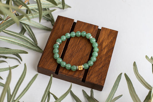 A bracelet of round green beads with a gold-coloured accent bead rests on a wooden block beside leaves.