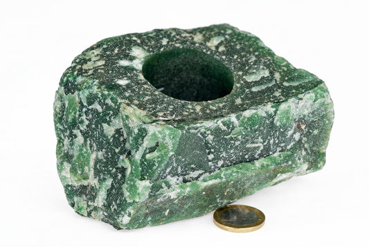 A green and white speckled stone object with a central hole, resting on a pale surface next to a coin.
