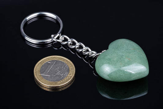 A silver-toned keychain with a heart-shaped green aventurine pendant and chain, next to a one-euro coin on a dark surface.