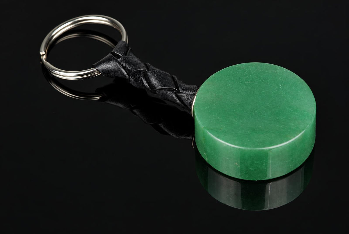A round green aventurine keychain with a braided black leather strap and silver-toned metal ring on a dark reflective surface.
