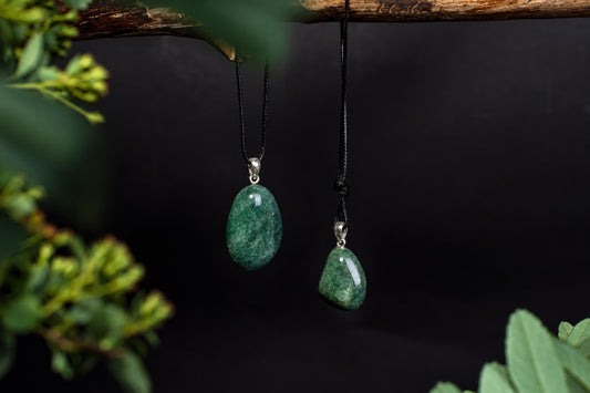 Two green aventurine pendants hang from black cords on a dark surface, framed by blurred green leaves.