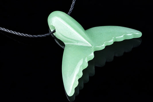 A light green pendant shaped like a dolphin tail hangs from a dark cord against a black reflective surface.