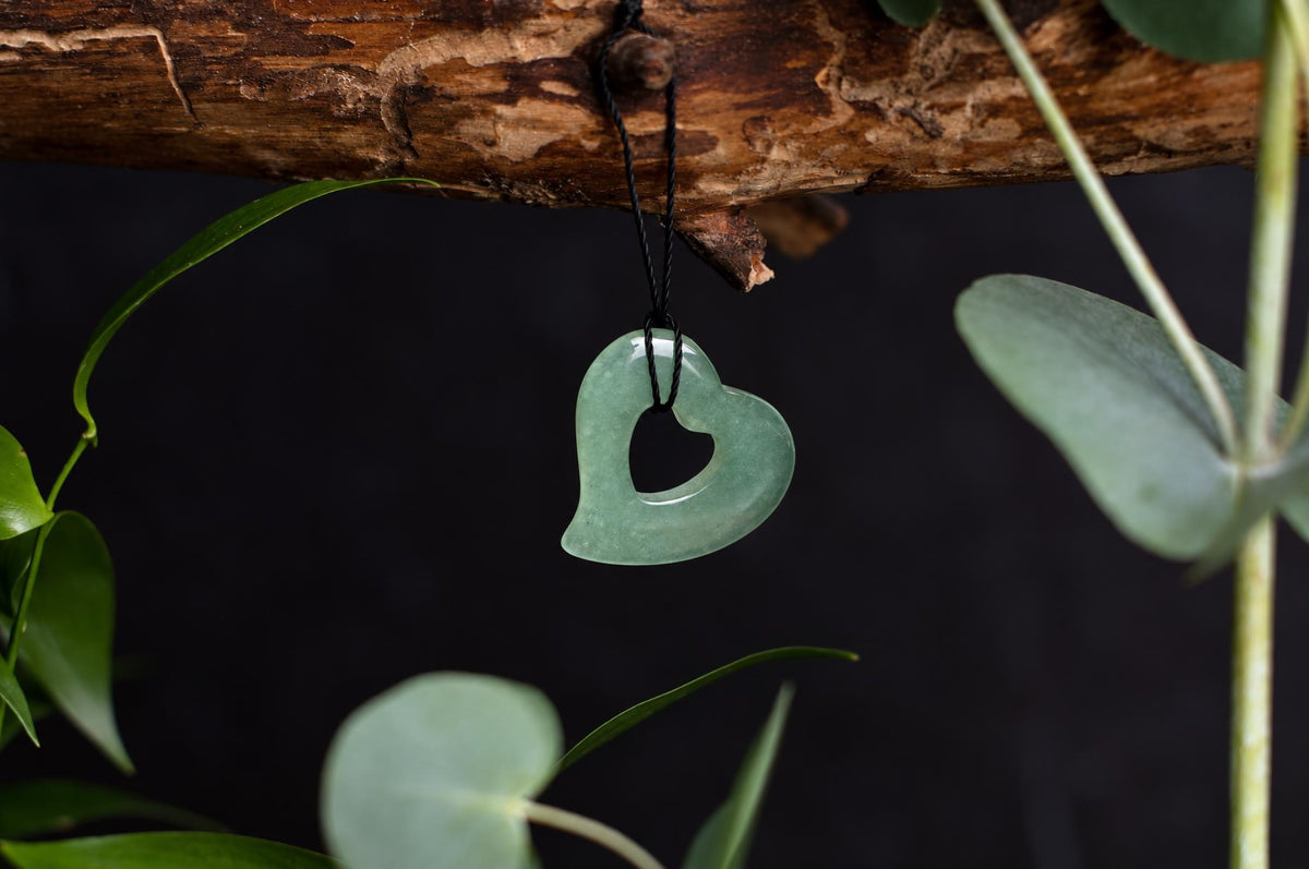 A green aventurine heart-shaped pendant with a central cutout hangs from a black cord on a wooden branch, surrounded by leaves.