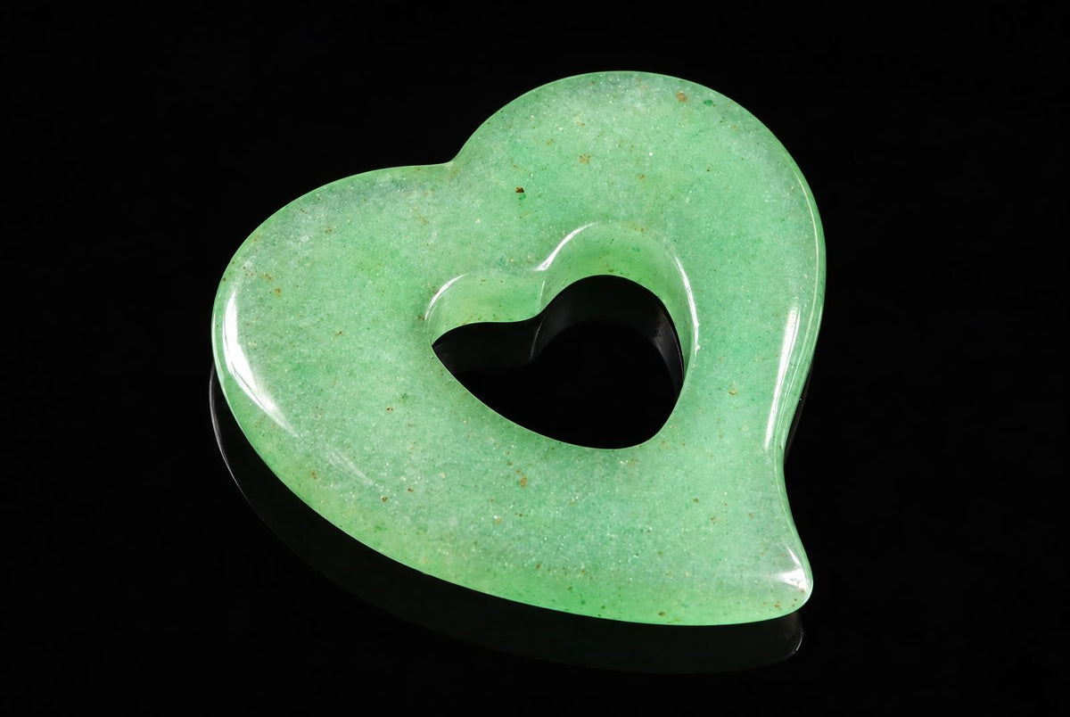 A polished, light green aventurine pendant shaped like a heart with a smaller heart-shaped cutout in the center, set against a dark surface.