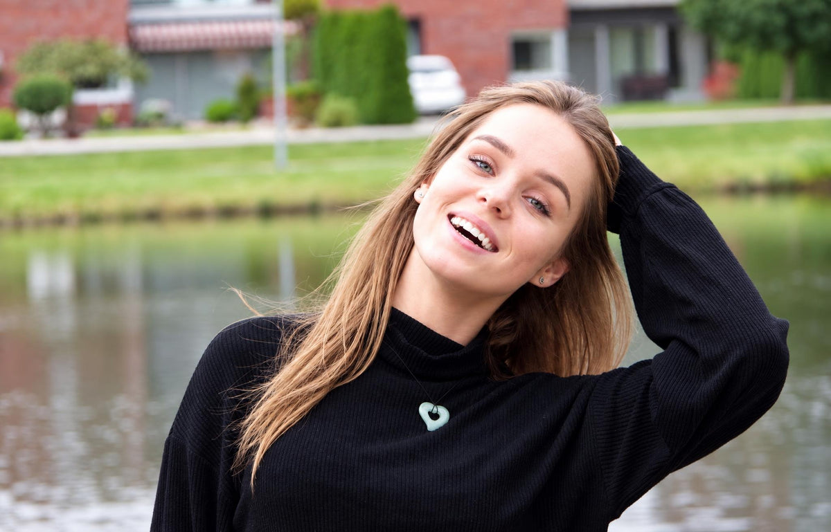 A smiling woman with long hair wears a black turtleneck and a light green heart-shaped pendant necklace, standing by water.