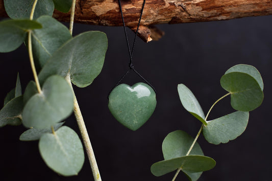 A polished green heart-shaped aventurine pendant hangs from a black cord beneath a wooden branch, surrounded by green leaves against a dark background.