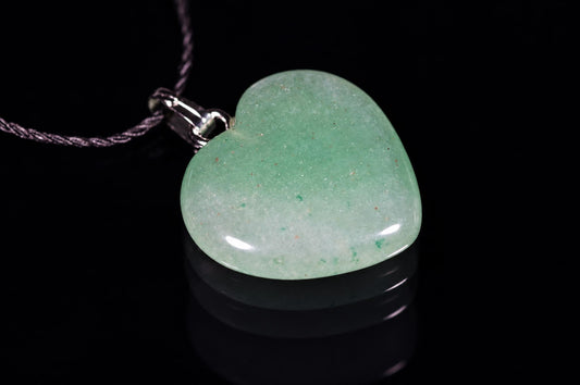 A polished heart-shaped aventurine pendant with a green, speckled surface hangs from a dark cord on a black reflective surface.