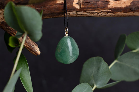 A polished green pebble-shaped pendant hangs from a dark cord beneath a wooden branch, surrounded by green leaves against a dark background.