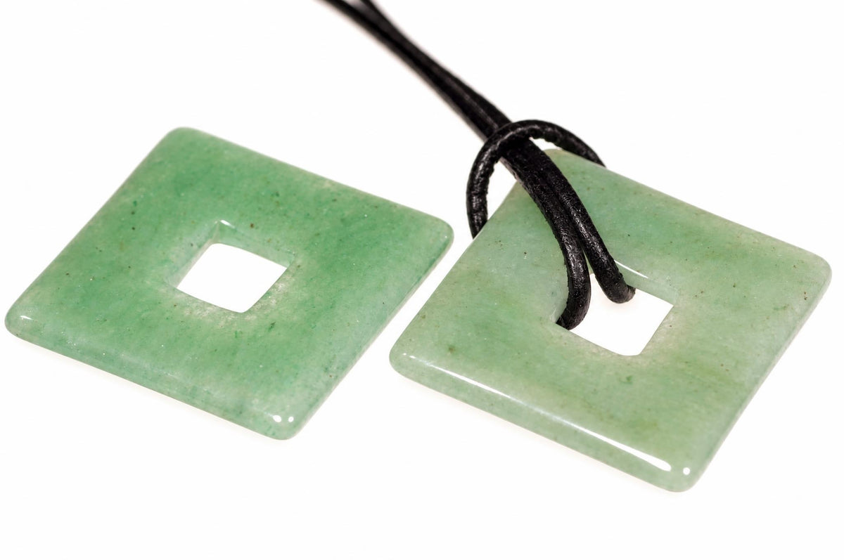 Two square, light green aventurine pendants with a central hole lie on a white surface; one has a black cord threaded through.