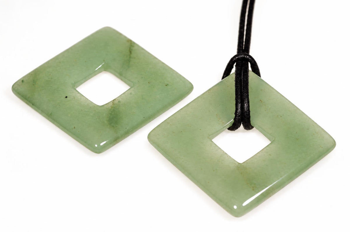 Two square, light green aventurine pendants with a central hole; one hangs from a black cord on a white surface.