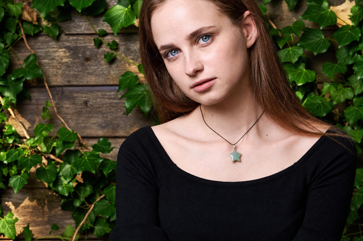 A woman wears a black top and a necklace with a pale green star pendant on a dark cord, against a wooden wall with ivy.