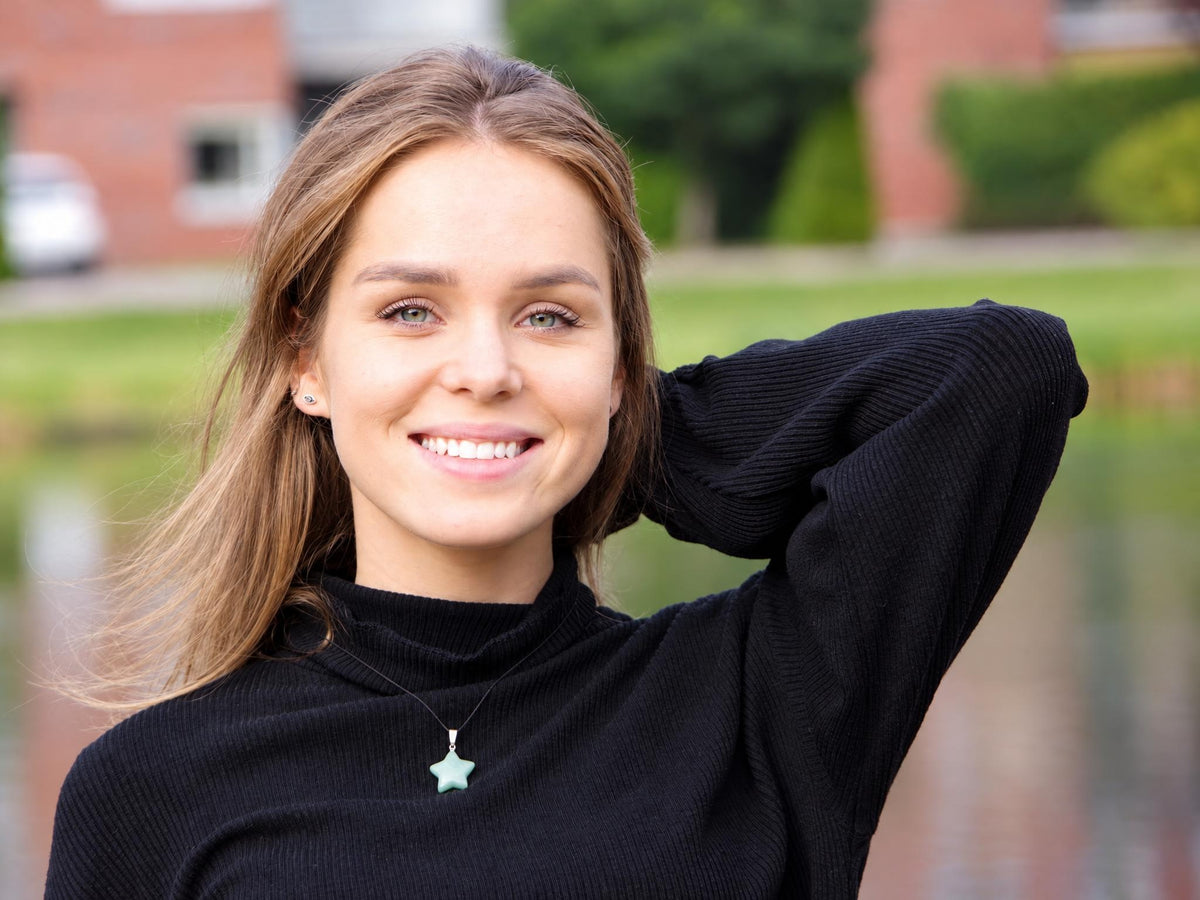 A woman wears a black turtleneck with a light green star-shaped pendant on a thin cord against a blurred outdoor background.