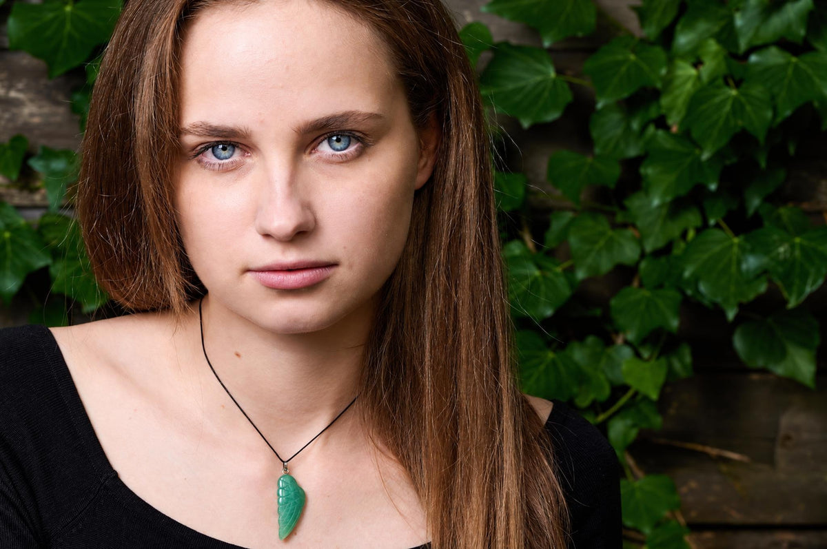 A woman wears a black top and a pendant with a green, wing-shaped stone on a dark cord against a leafy background.