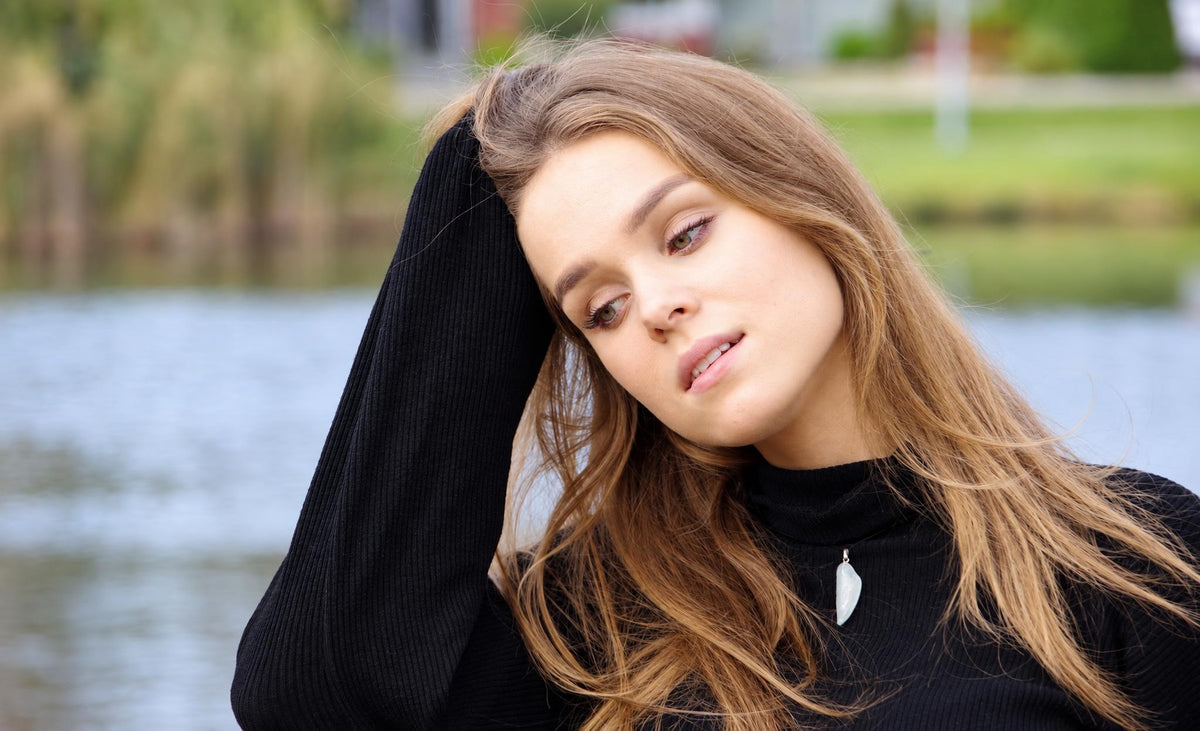 A woman with long brown hair wears a black turtleneck and a pale, wing-shaped pendant on a dark surface near water.
