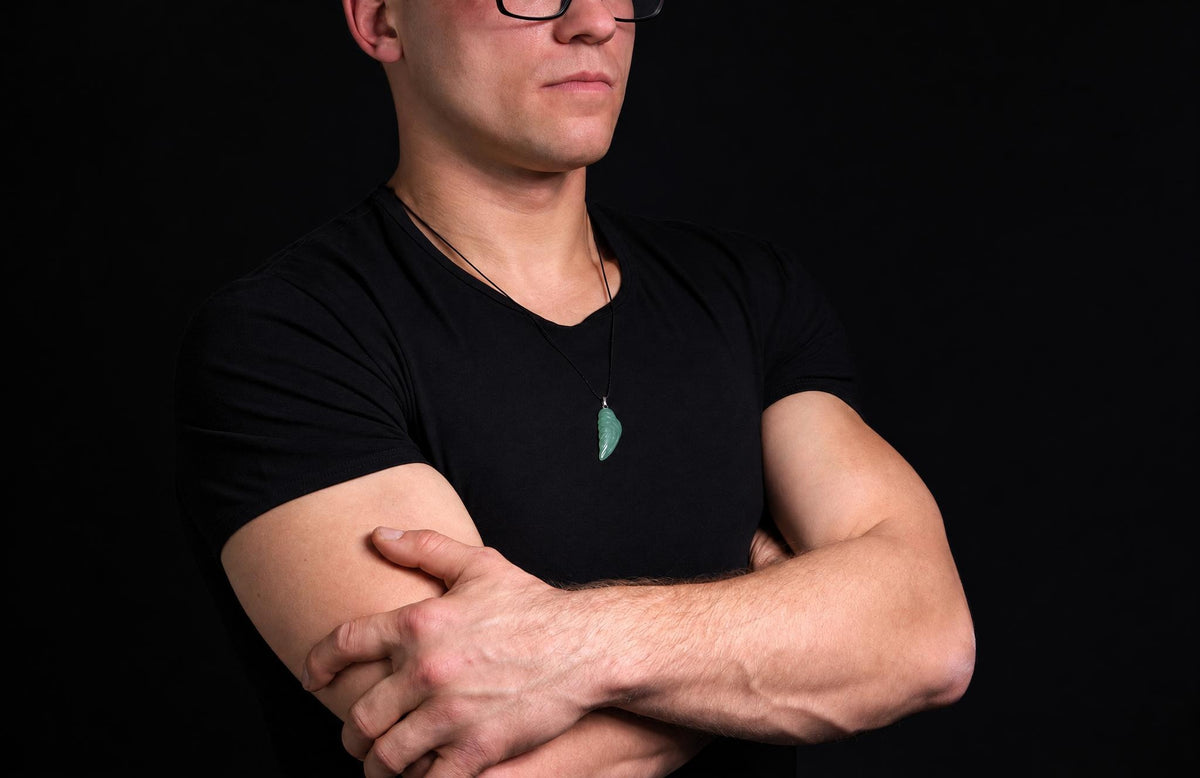 A man in a black t-shirt wears a green, wing-shaped pendant on a dark cord against a black background.
