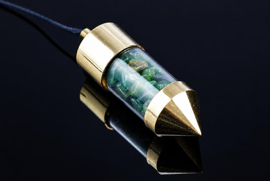 A brass pendulum with a clear tube containing green stones, attached to a dark cord, resting on a reflective black surface.