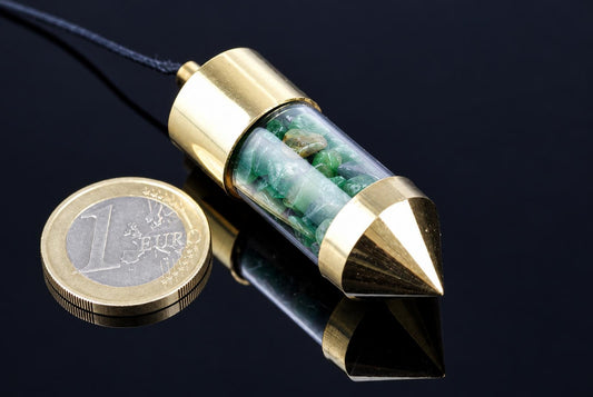 A brass pendulum with a clear tube containing green stones, attached to a dark cord, resting on a dark surface next to a one-euro coin.