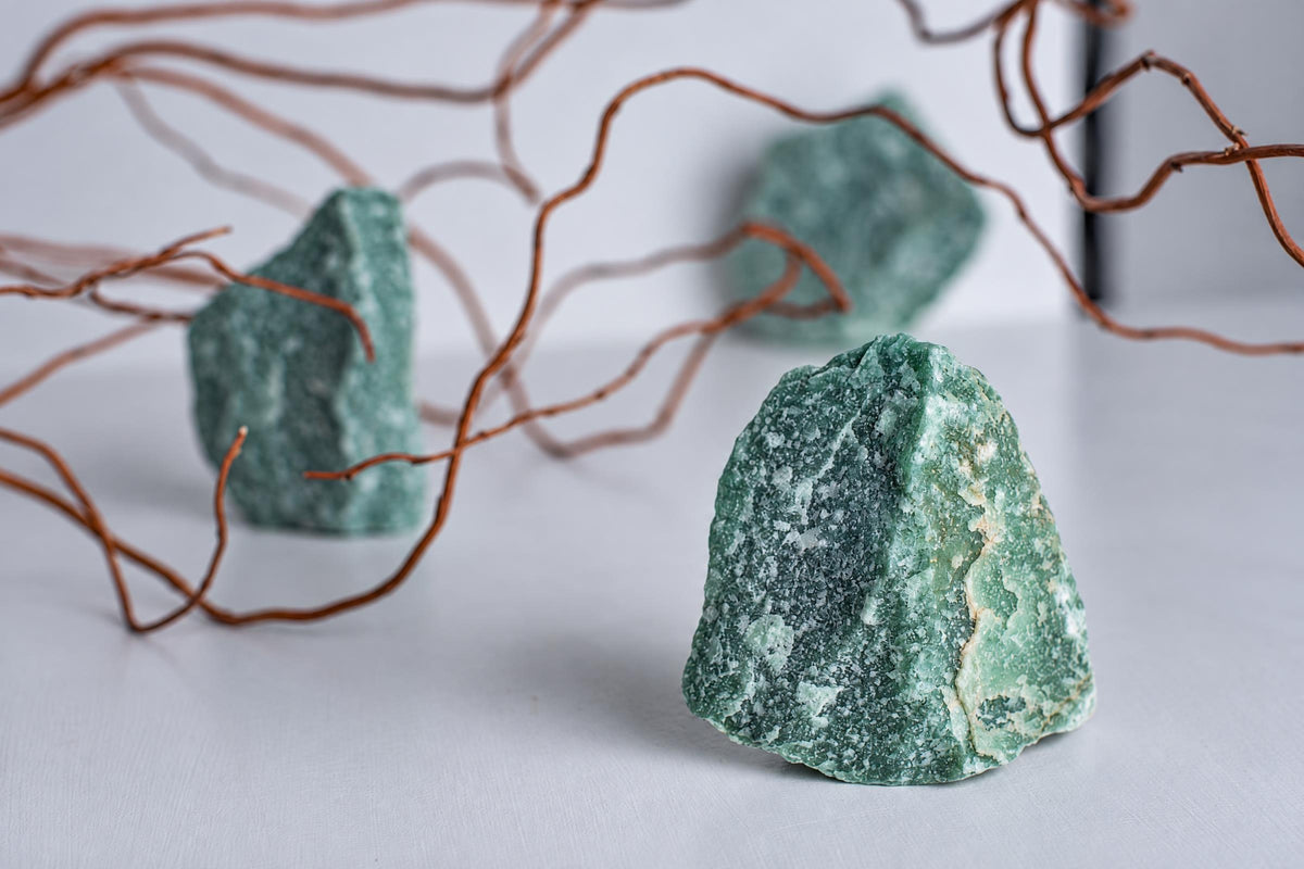 Three green aventurine stones with white speckles sit on a pale surface, framed by thin brown branches.