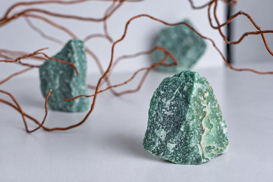 Three green aventurine stones with white speckles sit on a pale surface, framed by thin brown branches.