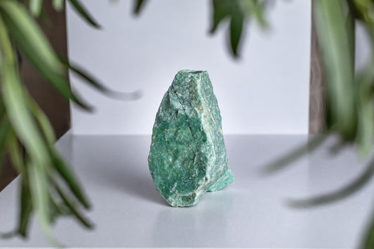 A green aventurine crystal stands on a white surface, framed by blurred plant leaves.