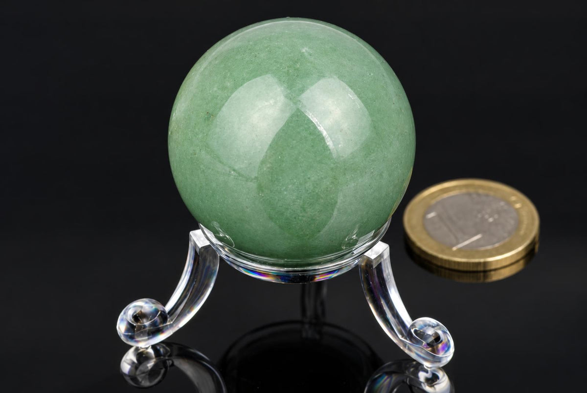 A polished green aventurine sphere rests on a clear acrylic stand against a dark reflective surface, with a coin nearby for scale.