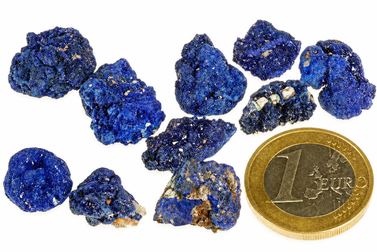 Several rough blue azurite mineral specimens with white and brown inclusions, arranged on a pale surface next to a one euro coin.
