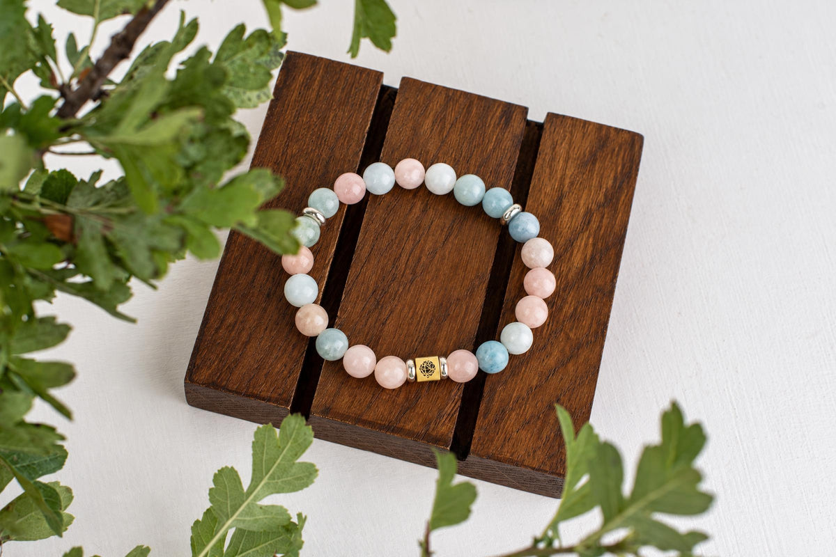 A beaded bracelet with pink, blue and white round beads on a dark wooden stand, surrounded by green leaves.