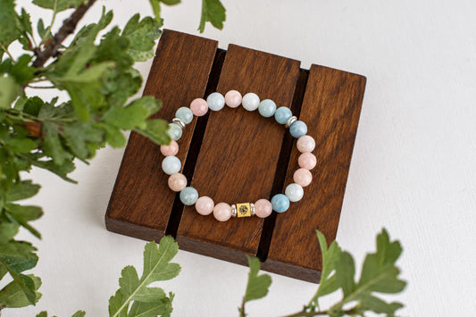 A beaded bracelet with pink, blue and white round beads on a dark wooden stand, surrounded by green leaves.