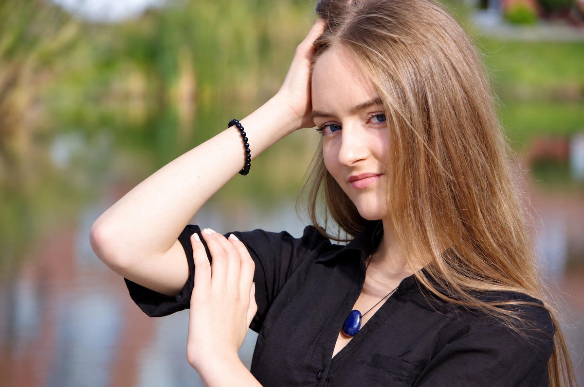 A woman wears a black beaded bracelet on her wrist and a blue oval pendant necklace against a blurred outdoor background.