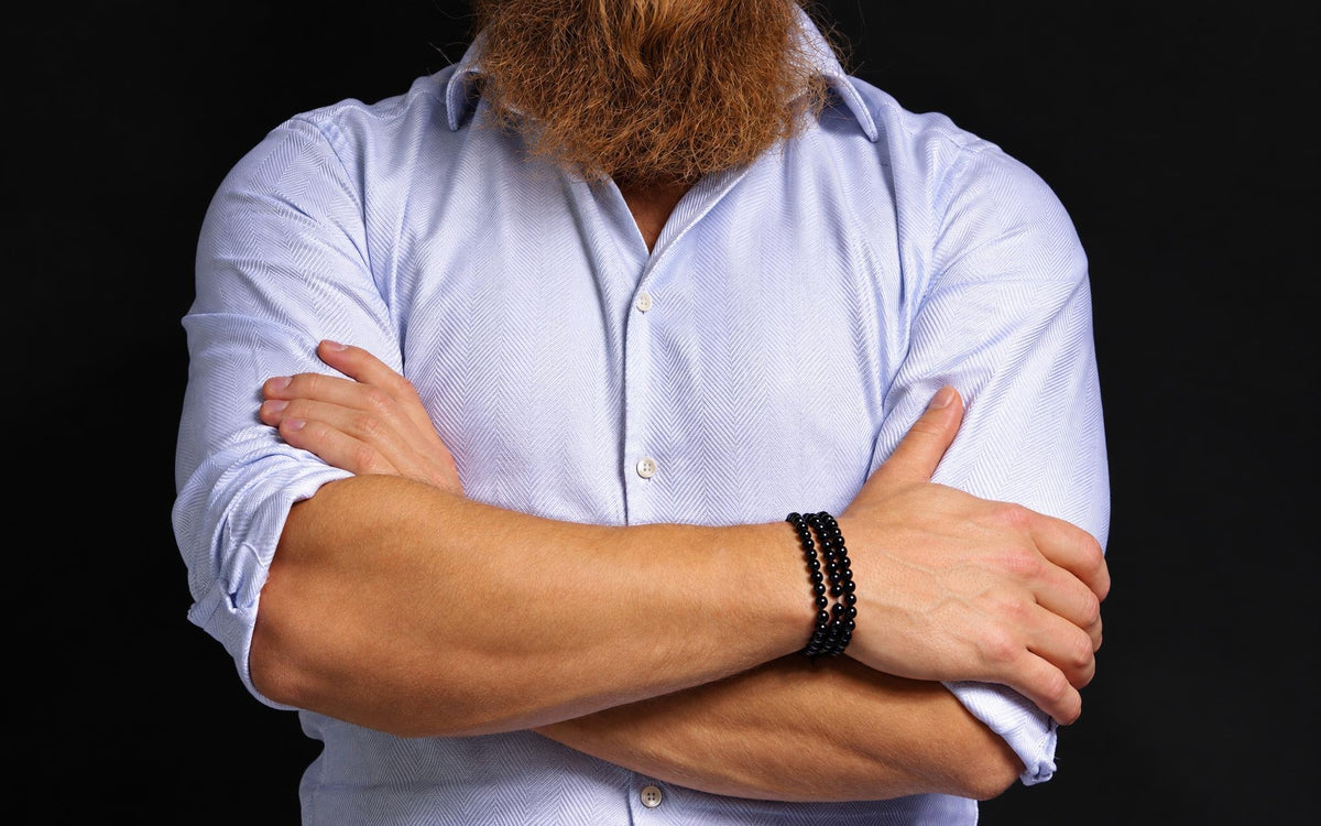 A man with a red beard wears a light blue shirt and three black beaded bracelets on his wrist against a dark background.